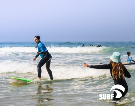 Apprenant debout sur une planche de surf sur la plage de Biscarrosse, devant une monitrice de GangSurf.